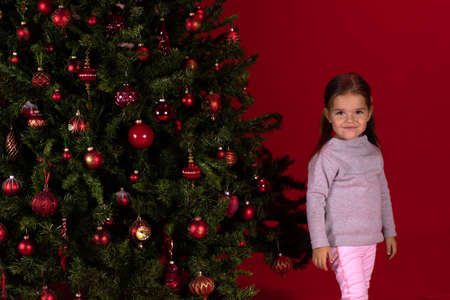 Portrait of little child girl next to Christmas tree on red studio Xmas backdrop. christmas, holidays and childhood concept. Merry Christmas! Happy New Year! Waiting for miracle concept.の写真素材