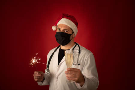 Doctor in protective mask and Santa Claus hat holds Burning sparklers and glass of champagne isolated on dark red studio copy space. Merry Christmas and Happy New Year for healthcare professionalsの写真素材