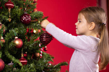 Focused happy toddler girl decorating Christmas tree hanging decorative toy, red studio Xmas background. Children and Christmas concept. preparation for holidays. Merry Christmas! Happy New Year!の写真素材