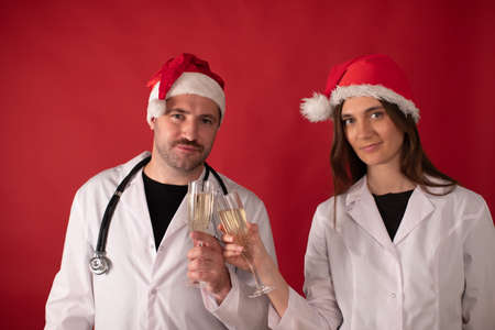 Two smiling doctors in Santa Claus hats cheering with champagne flutes looking at camera on red studio background. male and female Therapists Celebrating Christmas or New Year on duty.の写真素材