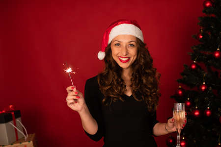 Beautiful long-hair brunette girl in Santa Claus hat sends Christmas greetings to her loved ones by zoom celebrating New Years Eve alone. Christmas tree on studio red backdrop copy space Festive moodの写真素材