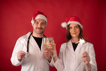 Two smiling doctors in Santa Claus hats cheering with champagne flutes and Burning sparklers looking at camera on red studio. Merry Christmas and Happy New Year for healthcare professionalsの写真素材