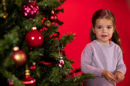 Smiling happy child baby girl posing at Christmas tree on red studio Xmas background. Children and Christmas concept. preparation for holidays. Merry Christmas! Happy New Year!の写真素材