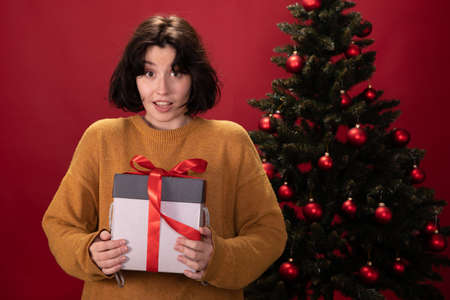 Young pretty woman looks surprised bewildered holding xmas gift box standing in front of Christmas...