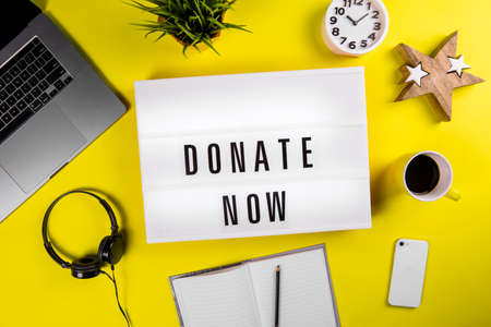 Donate Now message on lightbox on modern yellow office desktop with laptop, smartphone, coffee mug, notebook. call for donations, compassion, money transfers to charity organizations, philanthropyの写真素材