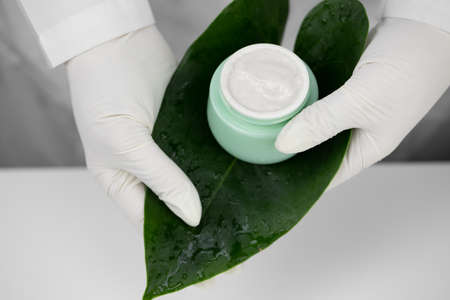 Close-up hands in medical gloves hold cream jar on green leaf, white backgroundの写真素材