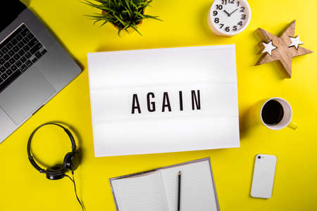 Lightbox with text message AGAIN on yellow background with laptop, smartphone, headphones, clock, notes. Concept of office workspace, business deadline, time to study, education, repetition learningの写真素材