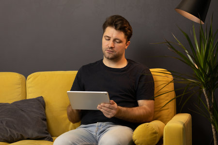Young unshaven handsome guy chatting and messaging tablet online sitting on sofaの写真素材