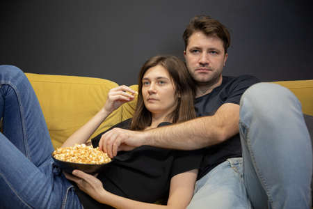 unemotional couple hugging lying on couch with popcorn in hands and watching TVの写真素材