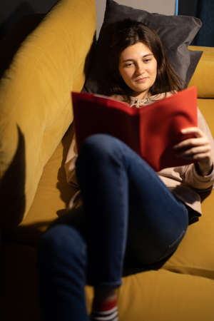 Attractive long-haired girl intently reads book lying on comfy sofa knees bentの写真素材