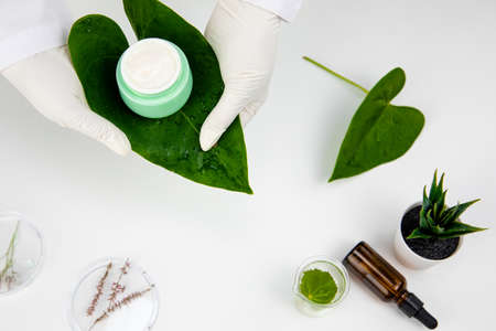 hands of female lab technician hold skincare cream in glass jar on tropical leafの写真素材