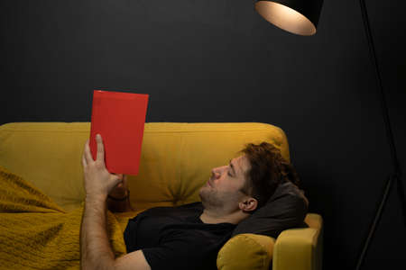 Young unshaven handsome guy reads book liyng on comfortable yellow sofaの写真素材