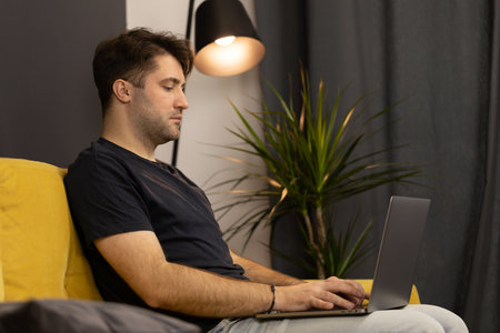 Young unshaven handsome guy man sitting on sofa and typing on laptop keyboardの写真素材