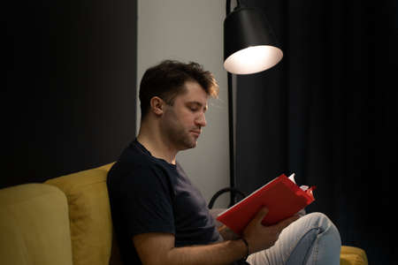 Young unshaven handsome guy reads book sitting on yellow sofa under spot lampの写真素材