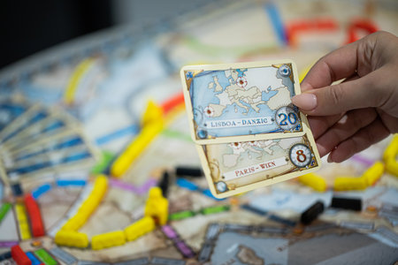 London, GB 11.02.2021 - Popular family railway-themed board game Ticket to Ride Europe edition. Player holding destination tickets in hand, map with colorful train cars and station out of focusのeditorial素材