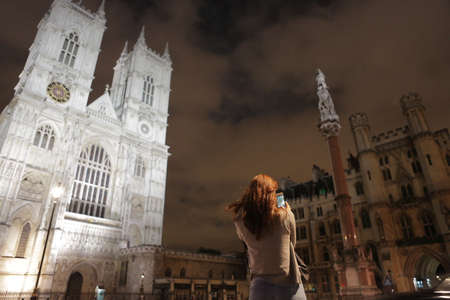 London, UK, 2.09.2019 - long-haired Redhead girl tourist takes smartphone photo of Westminster Abbey column. Collegiate Church of St Peter west facad at night. place of coronation of British monarchsのeditorial素材