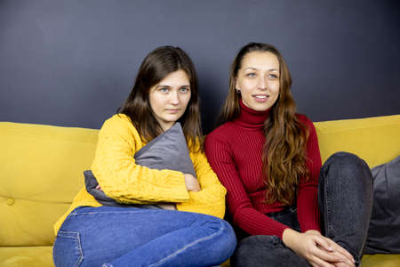 two pretty long-haired brunette girls sit on sofa watching TV huddled togetherの写真素材