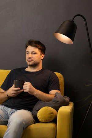 unshaven bored man with cup of coffee looking away sitting on comfy yellow sofaの写真素材
