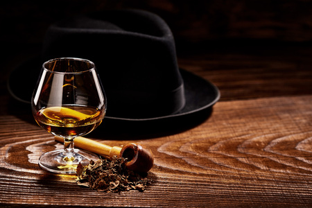 Glass of cognac and pipe with tobacco with black hat defocused on second background on wooden tableの写真素材