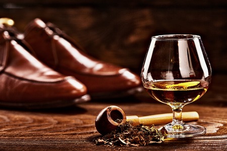 Glass of cognac and pipe with tobacco with glance shoes defocused on second background on wooden tableの写真素材