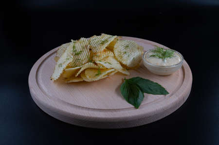 Potato chips with dill and sauce on a round wooden board. Isolated black background.