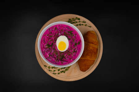 Summer cold beetrot soup (choldnik) with boiled egg served in a bowl on a wooden board with a croissant. Isolated black background. Top view.の写真素材