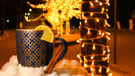A cup of tea with a slice of lemon and cinnamon in the snow on a winter night, with blurred lighted...