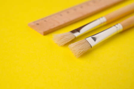 Close-up of paintbrushes next to a wooden ruler on a bright yellow background.