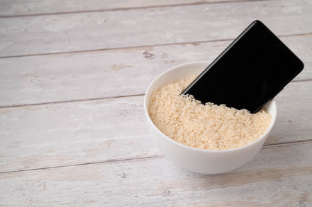 A mobile phone placed to dry in a bowl full of rice after the phone fell into water. Light wooden table background.の写真素材