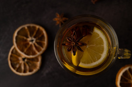 Close up of a glass teacup with black tea with lemon slices, star anise and cinnamon on a black...