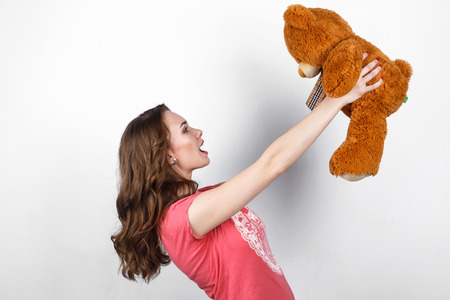 Stylish beautiful girl with long dark wavy hair in pink shirt holding in hands big brown teddy bear, gift in Valentine's Dayの写真素材
