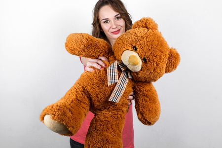 Stylish beautiful girl with long dark wavy hair in pink shirt holding in hands big brown teddy bear, gift in Valentine's Dayの写真素材