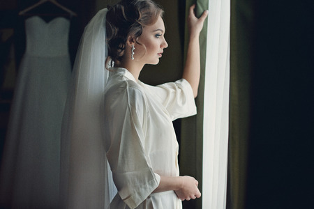 Beautiful bride in robe posing near window, wedding preparationの写真素材