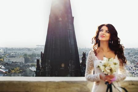 Bride daydreams with lily bouquet in her arms with cityscape on the backgroundの写真素材