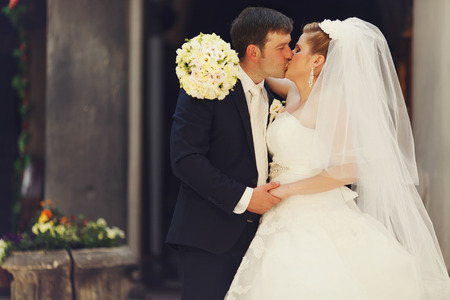 Bride holds a hand with bouquet on groom's shoulders while kissingの写真素材