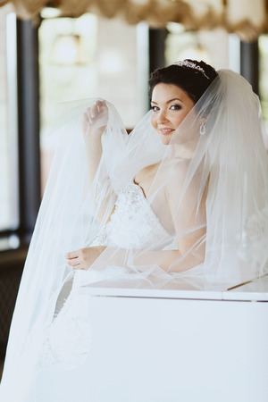 Bride smiles over a veil while she stands leaned on the pianoの写真素材