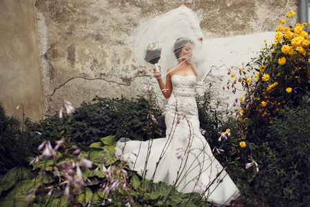 Wind blows bride's veil to an old wall while she stands among the flowersの写真素材
