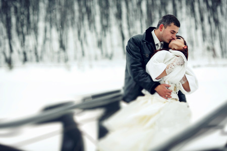 Groom kisses a bride in a white fur coat holding her in his arms in a winter dayの写真素材