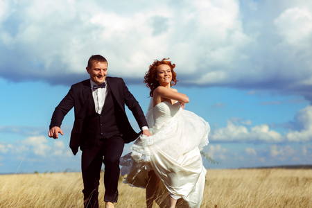 Bride rises her dress up walking across the field with a groom under clouded skyの写真素材
