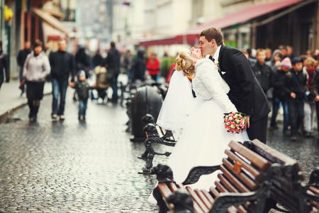 Groom kisses bride's neck standing between benches on a crowded streetの写真素材