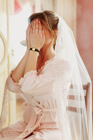 Bride in a veil sits on the chair and hides her face behind her palmsの写真素材