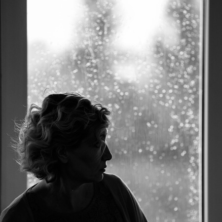 Woman with a curly hair sits thoughtful behind a window while rain fallsの写真素材