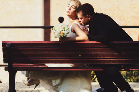 Groom kisses bride's shoulder while they rest in the sunrays on a benchの写真素材