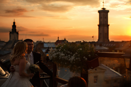 Groom holds bride's waist standing on the roof and wathcing a sunset over the cityの写真素材