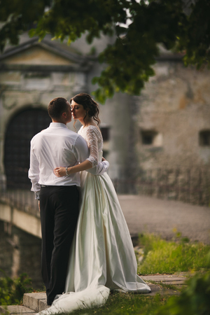 Groom hugs a brunette bride standing on the beginning of a way to an old castleの写真素材