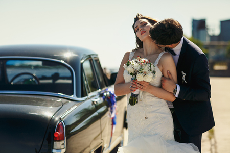 Stunning bride enjoys groom's kiss standing behind an old car in a windy weatherの写真素材