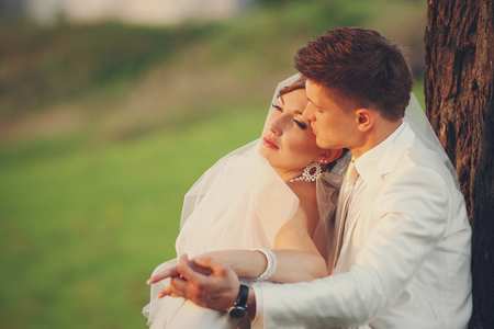 Gorgeous bride leans to groom's shoulder sitting in the rays of sunsetの写真素材