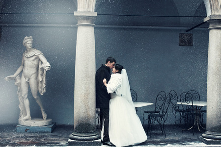 Picture in blue tones of newlyweds kissing behind a pillar while snow falls downの写真素材