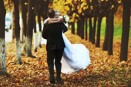 Happy bride holds groom's neck tightly while he whirls her on the fallen leavesの写真素材
