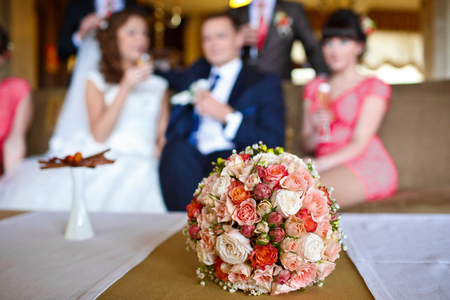 Pink and red wedding bouquet lies on the table in the front of newlyweds sitting on the coatの写真素材